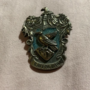 Ravenclaw emblem brosch - Snygg brosch med Ravenclaw-emblem från Harry Potter. Designen har en fågel och en trollkarlshatt i antik stil med en blå bakgrund. 