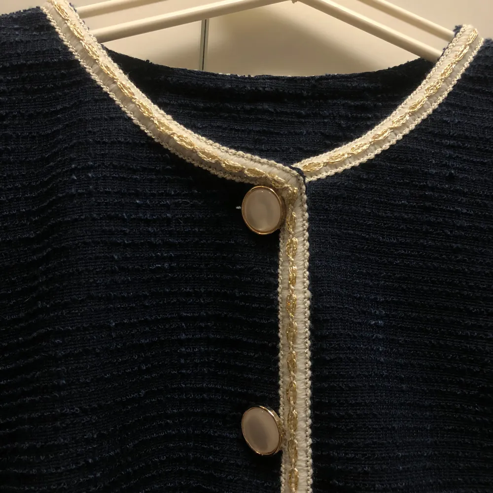 Elegant mörkblå kavaj med matchande kjol. Kavajen har långa ärmar och knäpps med stilrena knappar framtill. Både kavajen och kjolen har dekorativa guldfärgade kanter som ger en lyxig touch. Perfekt för en stilren look.. Puvut & Bleiserit.