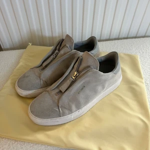 Grå sneakers från Axel Arigato - Snygga grå sneakers från Axel Arigato i mocka med dragkedja framtill. Skorna har en stilren design och är perfekta för en modern look. De kommer med en praktisk dustbag för förvaring. Använda fåtal gånger då dom är lite för små så säljer vidare till någon som får användning för dom🤩