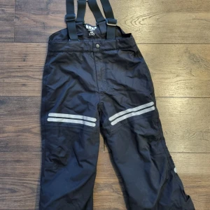 Svarta vinterbyxor med hängslen från Kaxs - Svarta vinterbyxor från Kaxs med justerbara och avtagbara hängslen och reflexdetaljer på benen. Snölås vid benslut. Justerbar midja. Perfekta för vinteraktiviteter med sitt slitstarka material och bekväma passform.