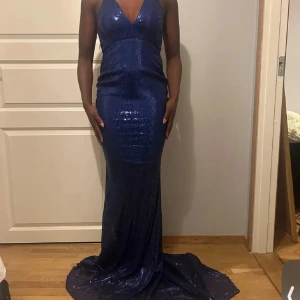 Blå glittrig långklänning som passar för er som har bal och mer sånna event ! - Snygg blå långklänning med glittrande paljetter och djup v-ringning. Klänningen har en öppen rygg med smala axelband som korsas. Perfekt för en glamorös kväll!