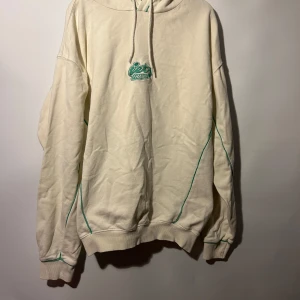 Beige hoodie från iets frans... - Vit hoodie med grön logotyp på bröstet och grön kant runt huvan och längs ärmarna. Ribbstickade muddar nedtill och vid ärmslut. Snörning i huvan. Storlek S. Hoodien har en flera fläckar