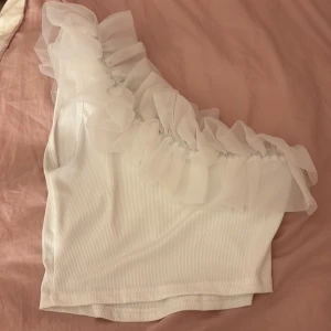 Vit oneshoulder topp med volang - Säljer en stilren vit oneshoulder topp med volangdetaljer. Toppen har en ribbad struktur och är perfekt för en trendig look. Passar bra till både jeans och kjol.