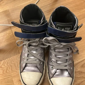 Grå och blå Converse All Star sneakers - Säljer ett par grå och blå Converse All Star sneakers med hög modell och snörning. Skorna har en klassisk design med vita sulor och Converse-logotypen på sidan. Perfekta för en avslappnad stil.