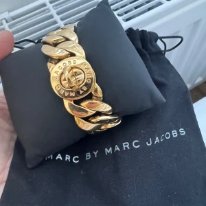 Guldarmband från Marc by Marc Jacobs - Snyggt guldarmband från Marc by Marc Jacobs med breda länkar och en rund detalj med logotyp. Perfekt för att ge en lyxig touch till din outfit. Kommer med en svart förvaringspåse.