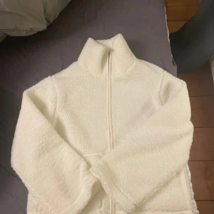 Vit teddyjacka med dragkedja - Mysig vit teddyjacka med dragkedja framtill. Jackan har en hög krage och långa ärmar, perfekt för kyliga dagar. Den fluffiga texturen ger en trendig look och extra värme.