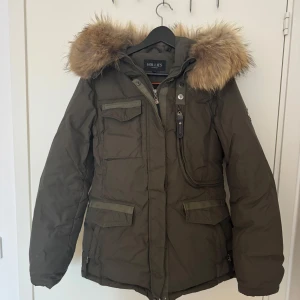 Olivgrön parkajacka med pälsdetaljer från Hollies - Snygg olivgrön parkajacka från Hollies med avtagbar huva och pälsdetaljer. Jackan har flera praktiska fickor och dragkedja framtill. Perfekt för kyliga dagar med sin varma och skyddande design.