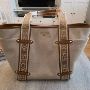 Beige shoppingväska från Michael Kors - Snygg beige shoppingväska från Michael Kors med bruna detaljer och logotypband. Väskan har en rymlig insida med dragkedjeficka och tryckknappsstängning. Perfekt för att bära med sig allt man behöver under dagen. Skickas med original dust bag