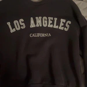 Svart sweatshirt från Gina Tricot - Säljer en svart sweatshirt från Gina Tricot med texten 'Los Angeles California' tryckt på framsidan. Tröjan har en klassisk rund hals och långa ärmar. Perfekt för en avslappnad stil.