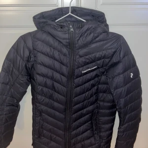 Svart dunjacka från Peak Performance - Säljer en stilren svart dunjacka från Peak Performance. Jackan har en dragkedja framtill och en praktisk huva.