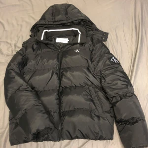Svart pufferjacka från Calvin Klein Jeans - Säljer en svart pufferjacka från Calvin Klein Jeans med huva och dragkedja. Jackan har CK-logga på bröstet och ärmen. Perfekt för kyliga dagar.