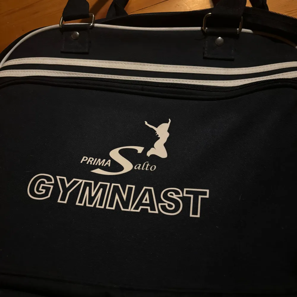 Säljer en svart gymväska från Prima Salto med texten 'GYMNAST' tryckt på framsidan. Väskan har dubbla handtag och en dragkedja upptill. Perfekt för att ta med till träningen eller gymmet!. Väskor.