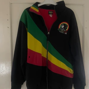 Svart reggae-jacka med Bob Marley-motiv - Säljer en svart jacka med reggae-vibbar och ett coolt Bob Marley-motiv på bröstet. Jackan har en dragkedja framtill och färgglada ränder i grönt, gult och rött som går diagonalt över både fram- och baksidan. Insidan är röd vilket ger en snygg kontrast. Perfekt för dig som vill ha en unik och avslappnad stil!