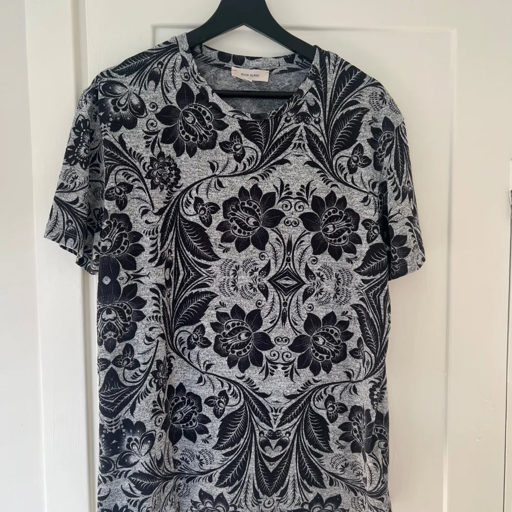 Säljer en snygg t-shirt från River Island i storlek M. Den har ett coolt svart och grått blommigt mönster som verkligen sticker ut. Perfekt för både vardag och fest! T-shirten är kortärmad och gjord i ett mjukt material som känns skönt mot huden.. T-shirts.