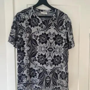 Säljer en snygg t-shirt från River Island i storlek M. Den har ett coolt svart och grått blommigt mönster som verkligen sticker ut. Perfekt för både vardag och fest! T-shirten är kortärmad och gjord i ett mjukt material som känns skönt mot huden.