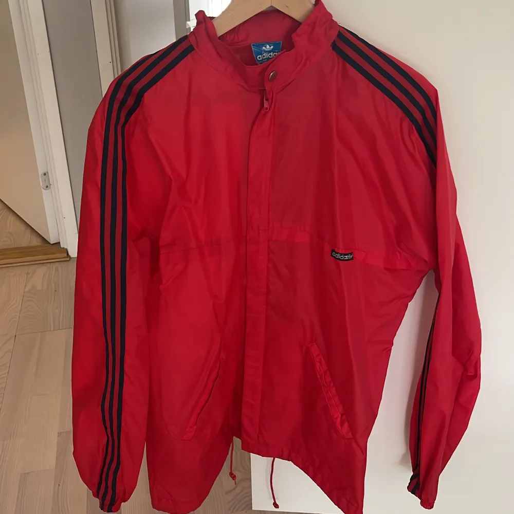 Säljer en snygg röd vindjacka från Adidas med klassiska svarta ränder längs ärmarna. Jackan har en dragkedja framtill och är tillverkad i 100% polyamid, vilket gör den lätt och perfekt för blåsiga dagar. Den har en normal passform och är långärmad. Perfekt för vår och höst!. Jackor.