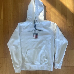 Vit hoodie från GANT - Säljer en snygg vit hoodie från GANT i storlek M. Den har en klassisk design med GANT-loggan broderad på bröstet och en cool sköld med stjärnor och ränder. Hoodien har en känguruficka och justerbar dragsko i huvan. Perfekt för en avslappnad stil!