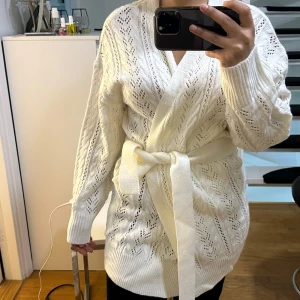 Stickad kofta  - En vit stickad kofta med hål och band runt midjan, helt oanvänd 