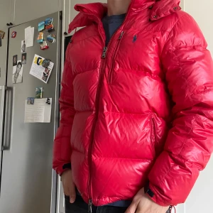 Ralph Lauren Jacka - Säljer denna snygga ralph lauren pufferjackan i röd färg med blått broderat märke. Den är använd endast en vinter och är alltså i väldigt fint skick. Storleken är M och passar som det också. Nypris 6000 kr. Kom privat för fler frågor!