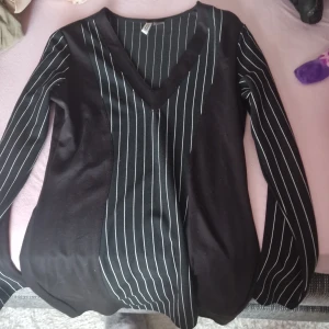 Svart v-ringad tröja från H&M - Säljer en snygg svart klänning från H&M:s Divided-kollektion. Den har ett stilrent vitt randigt mönster och en v-ringad hals. Perfekt för både vardag och fest! Klännimgen är långärmad och har en normal passform.