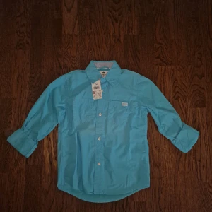 Turkos skjorta från KappAhl - Säljer en snygg turkos skjorta från KappAhl i storlek 134. Skjortan är långärmad och har en klassisk krage med knappar framtill. Perfekt för både vardag och festliga tillfällen. Den är helt ny med prislappen kvar. Inköpt för 129:- men jag säljer för