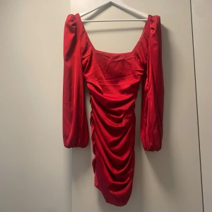 Röd klänning med rynkdetaljer - Säljer en superfin röd klänning med långa ärmar och rynkdetaljer framtill. Den har en tight passform och är perfekt för festliga tillfällen. Klänningen är kort och har en djup urringning som ger en elegant look. Materialet ser ut att vara glansigt, vilket ger en lyxig känsla. Använd endast 1 gång