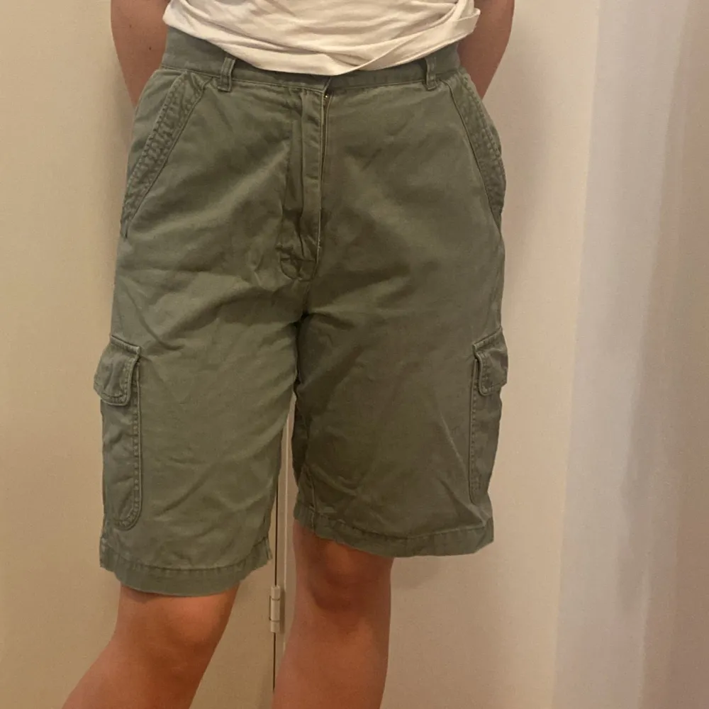 Säljer ett par gröna cargo shorts i bomull. De har flera praktiska fickor med knappar och en bekväm passform. Perfekta för sommarens äventyr eller en avslappnad dag ute. Shortsens stil är både funktionell och trendig. Uppskattad storlek M. Fattas en knapp på fickan.. Shorts.