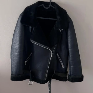 Svart skinnjacka med bälte - Säljer en snygg svart skinnjacka med bälte och dragkedja. Jackan har en cool bikerstil med långa ärmar och är perfekt för höst och vinter. Den har också praktiska fickor med dragkedjor. Perfekt för att ge din outfit en edgy touch! Knappt använd. Den är oversized