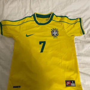 Ronaldinho Brasil-tröja från 1998! 🇧🇷🌟 - Säljer en ikonisk Ronaldinho Brasil-tröja från 1998 (replika)! Tröjan är som ny, bara provad en gång då storleken tyvärr inte passade mig. Perfekt för samlare eller för dig som vill ha en klassisk fotbollströja med en av tidernas bästa spelare. 