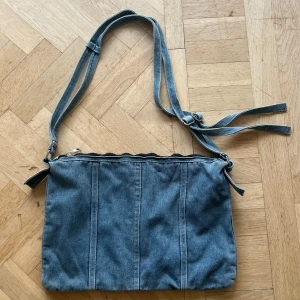 Blå axelväska i jeansmaterial - Säljer en snygg blå axelväska i jeansmaterial. Väskan har en justerbar axelrem och stängs med dragkedja. Perfekt för vardagsbruk och rymmer det mesta du behöver ha med dig. Den är i bra skick och redo för en ny ägare!