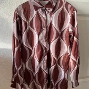 Brun och vit mönstrad skjorta - Säljer en snygg brun och vit skjorta med ett vågigt mönster. Skjortan har långa ärmar och knappar framtill. Perfekt för en stilren look, kanske till en fest eller en utekväll. Materialet känns bekvämt och luftigt.