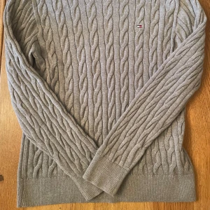 Tommy Hilfiger Stickad Grå Tjocktröja - En tvär fett stickad grå tjocktröja från Tommy hilfiger använd en gång, säljer för den passar inte mig. 10/10 skick original pris 1000 kr perfect för kallt väder som det kommer bli i vintern.  Pris kan diskuteras