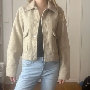 Beige jacka med knappar - Säljer en snygg beige jacka i normal passform. Den har långa ärmar och stängs med knappar framtill. Perfekt för höst och vår, och den har praktiska fickor med lock. Jackan är i nyskick och passar till både vardag och lite finare tillfällen.