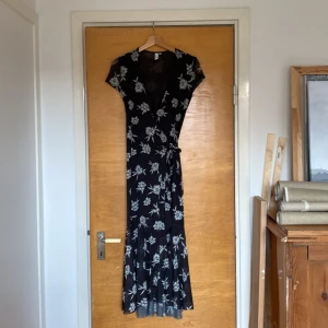 Mörkblå blommig omlottklänning  - En mörkblå (nästan svart) lång omlottklänning med korta ärmar och vitblommigt mönster. Följsam passform.