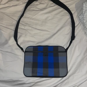 Burberry väska - Säljer en snygg burberry axelväska med ett rutigt mönster i blått och grått. Väskan har en justerbar axelrem och en dragkedja upptill. Perfekt för både vardag och fest! 👜