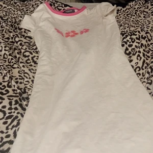 Vit klänning med rosa detaljer - Säljer en söt vit klänning med rosa kanter och blommigt mönster framtill. Klänningen är kortärmad och perfekt för sommardagar. Den är i bra skick och passar utmärkt till både vardag och festliga tillfällen.