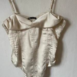 Beige korsett från Zara - Snygg beige korsett från Zara i ett glansigt satinmaterial. Den har smala axelband och en elegant krage som ger en lyxig känsla. Perfekt för festliga tillfällen eller en utekväll. Passar bra till både kjol och byxor!