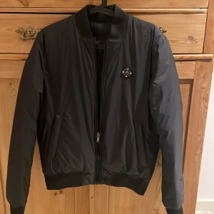 Svart bomberjacka - Säljer en snygg svart bomberjacka i mycket bra skick. Jackan har en dragkedja framtill och är quiltad för extra värme. Perfekt för höst och vår! Den har långa ärmar och en stilren design som passar till det mesta. Ett måste i garderoben för en cool och avslappnad stil, har två sidor en utan j.linderberg logga och en annan sida med logga ny pris 3000kr. 