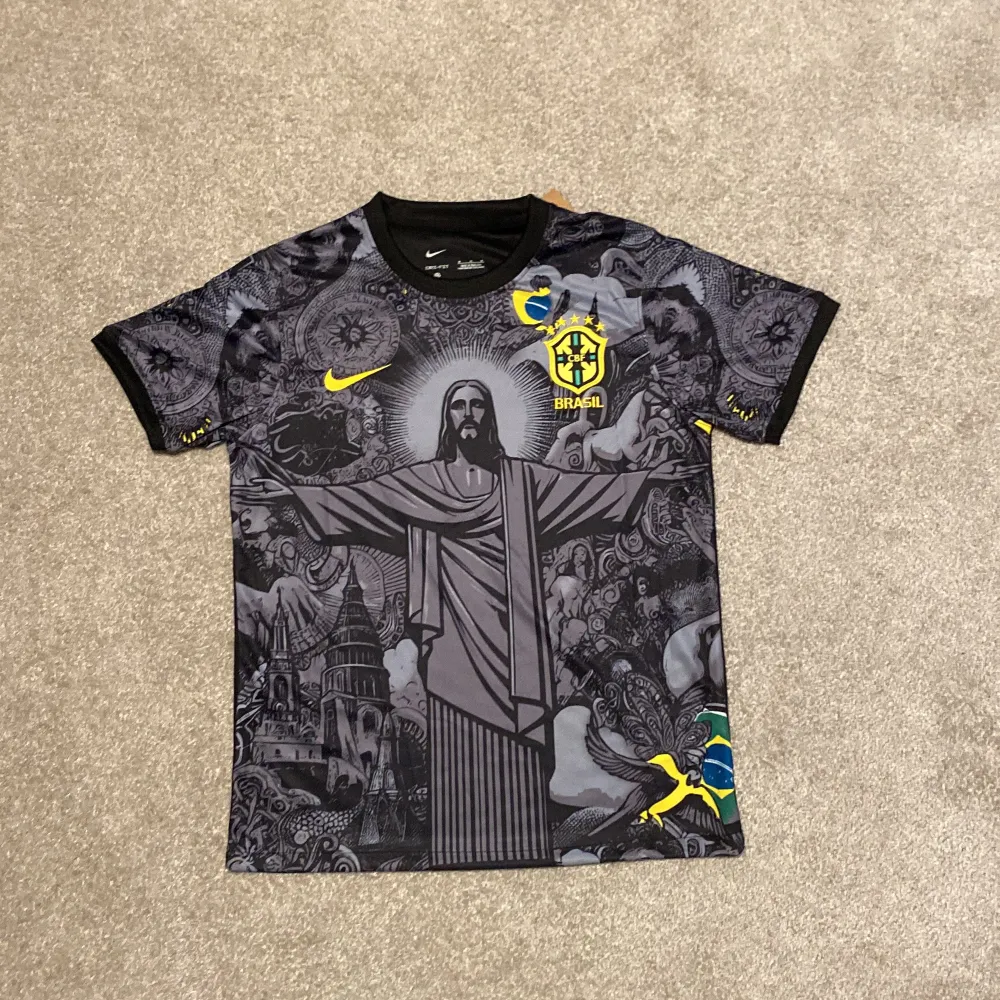 Säljer en grå fotbollströja från Nike med ett coolt Brasilienmotiv. Tröjan har korta ärmar och är i nytt skick. Den har ett detaljerat mönster med Kristusstatyn och andra symboler. Perfekt för fotbollsfans som vill sticka ut!. T-shirts.