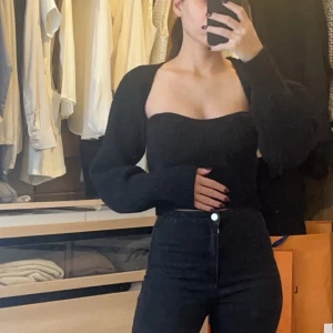 Svart axelbandslös topp med matchande bolero  - Snygg svart axelbandslös topp med en matchande bolero som passar perfekt för en utekväll eller fest. Den har en tight passform som framhäver figuren och är gjord i ett mjukt material för extra komfort. Perfekt att matcha med höga jeans eller kjol för en stilren look.