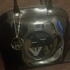 Svart handväska från Armani Jeans - Snygg svart handväska från Armani Jeans i glansigt syntetmaterial. Väskan har ett stort logotypmärke på framsidan och en praktisk dragkedja upptill. Insidan är fodrad med mönstrat tyg och har en innerficka med dragkedja. Perfekt för både vardag och fest!