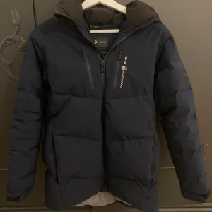 Mörkblå dunjacka från Sail Racing - Säljer en snygg mörkblå dunjacka från Sail Racing. Jackan har en praktisk dragkedja och en skön huva. Perfekt för kalla vinterdagar. Den är lite sliten Storlek 170 men passar mig som är 175 ny pris 4500kr 