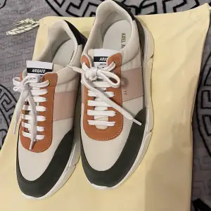 Säljer ett par snygga sneakers från Axel Arigato i nytt skick. Skorna har en cool färgkombination av vit, beige, grön och orange med snörning. Perfekta för både vardag och fest! De är gjorda i syntetmaterial och har en modern design som verkligen sticker ut.