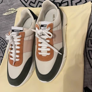 Sneakers från Axel Arigato - Säljer ett par snygga sneakers från Axel Arigato i nytt skick. Skorna har en cool färgkombination av vit, beige, grön och orange med snörning. Perfekta för både vardag och fest! De är gjorda i syntetmaterial och har en modern design som verkligen sticker ut.
