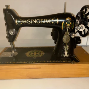 Antik symaskin från Singer - Säljer en vacker antik symaskin från Singer med serienummer H5757609. Den har en elegant svart finish med detaljerade blommönster i guld, rött och grönt. Maskinen är monterad på en träbas och har en klassisk handvev. Perfekt för samlare eller som en unik inredningsdetalj!