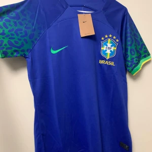 Blå Brasilien fotbollströja från Nike - Säljer en blå Brasilien fotbollströja från Nike i nytt skick. Tröjan har korta ärmar med grönt och gult leopardmönster. Den är slim fit och tillverkad i Brasilien. Perfekt för fotbollsfans som vill visa sitt stöd för laget! 💙💚💛