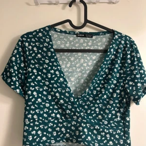 Blommig grön croppad topp från Shein - Säljer en superfin grön croppad topp med ett sött blommönster Toppen har en v-ringad design och korta ärmar, Den är i bra skick och redo för nya äventyr!💚