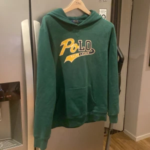 Grön hoodie från Ralph Lauren - Säljer en snygg grön hoodie från Ralph Lauren i storlek S-M. Den har en stor POLO-logga i gult och vitt på framsidan. Perfekt för höst och vinter med sin mysiga och varma känsla. Den har en klassisk känguruficka och ribbade muddar vid ärmslut och nederkant. Passar både till vardags och chillkvällar. 🧥