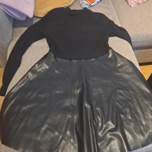 Svart klänning med ribbad överdel och skinnkjol - Snygg svart klänning med en ribbad överdel och en glansig skinnkjol. Perfekt för höst och vinter, med långa ärmar som håller dig varm. Klänningen har en tight passform upptill och en utsvängd kjol som ger en fin silhuett. Passar både till vardags och fest!