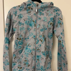 Grå hoodie med blommigt mönster - Grå kofta med blommigt mönster i turkos och grönt. Den har en dragkedja framtill och en huva. Den är i mycket bra skick men kommer ej till användning❤️